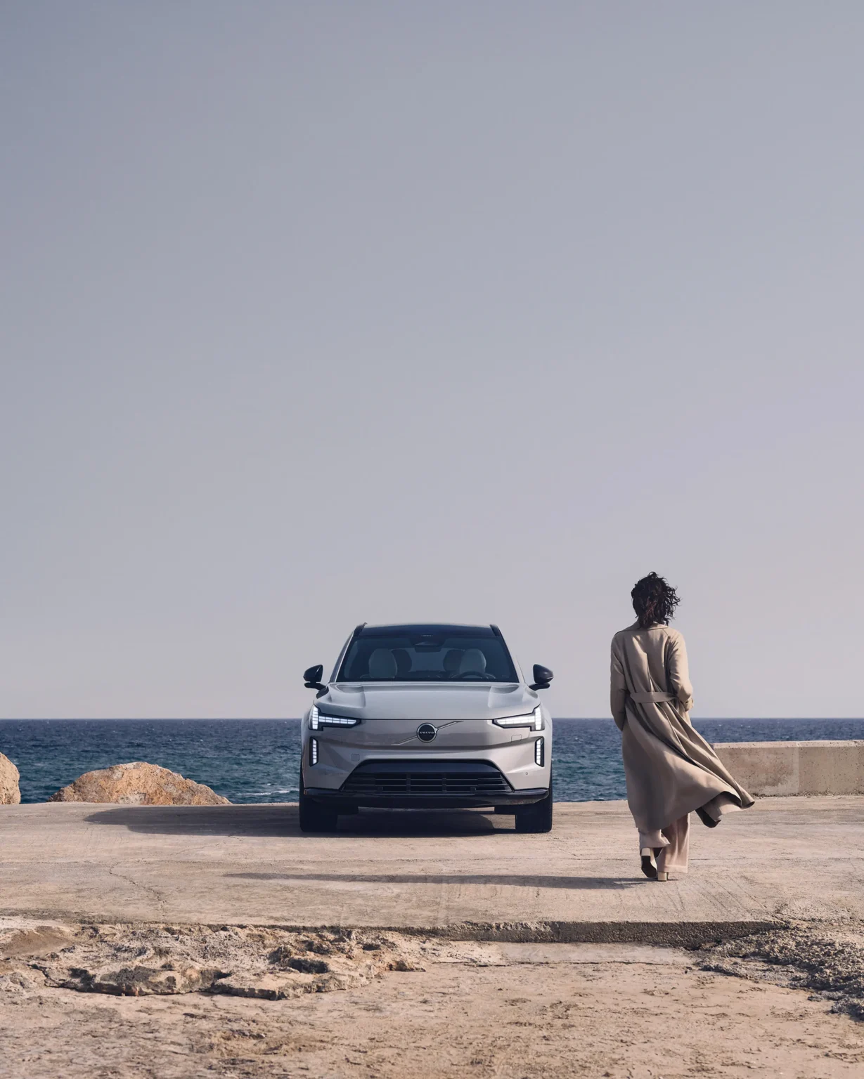 Volvo EX90 parked on a rocky seafront as a woman in a long coat walks past