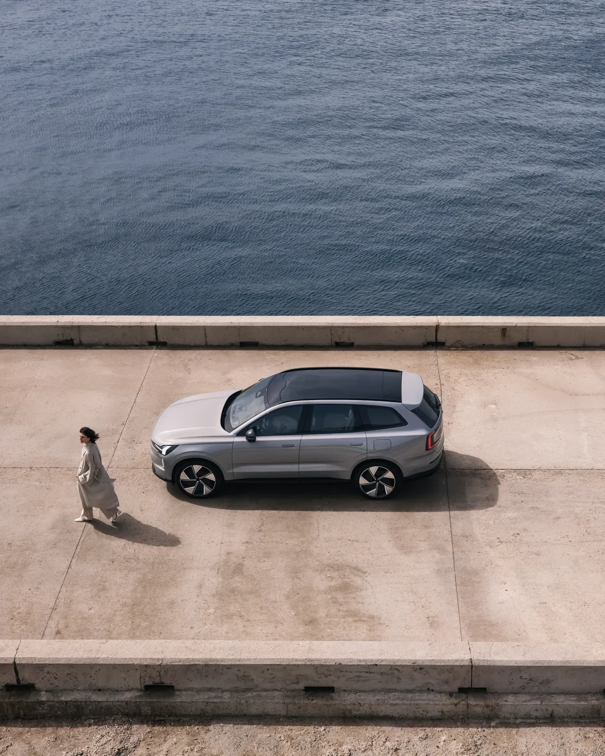 Aerial view of a Volvo EX90 parked on a concrete quay beside the sea, with a woman walking alongside