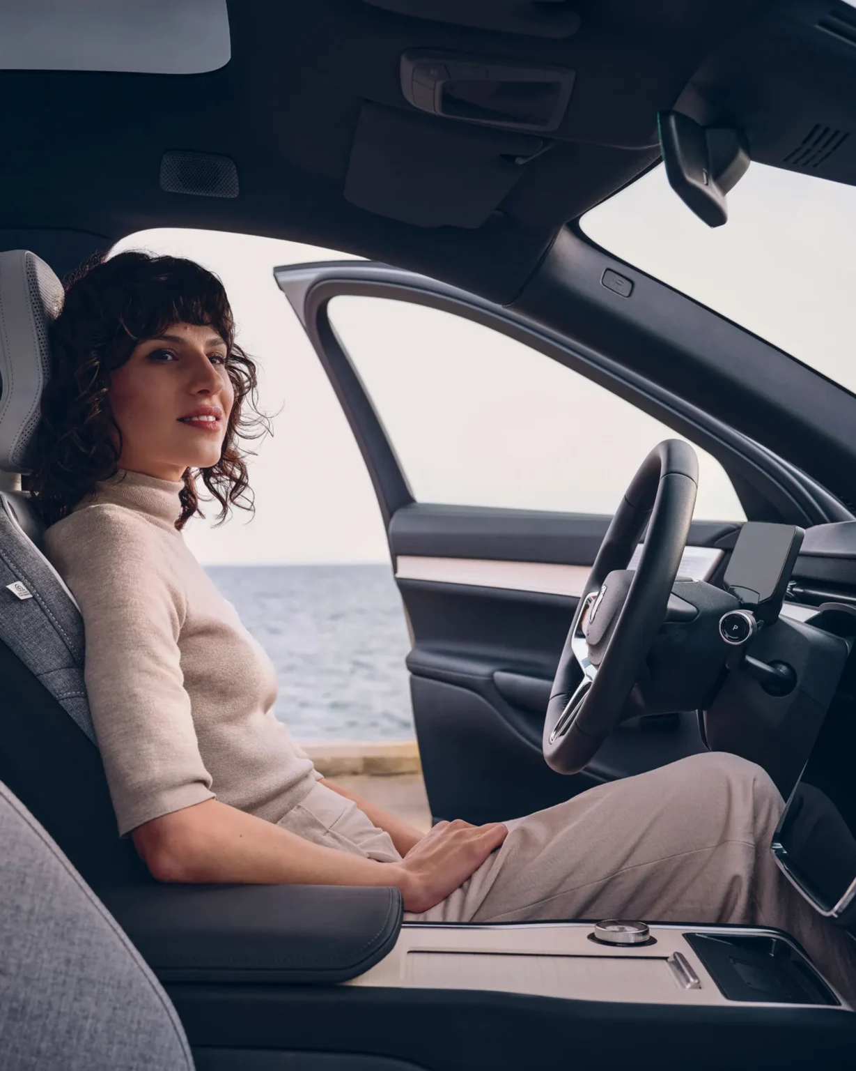 Woman sitting in the driver's seat of a Volvo EX90 with the door open, looking out towards the sea