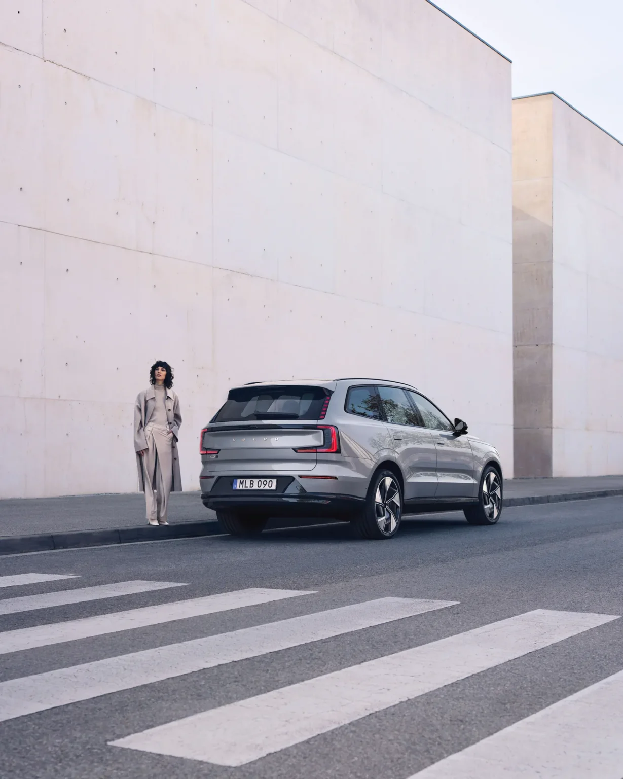 Woman standing beside a Volvo EX90 parked at a pedestrian crossing below tall white walls