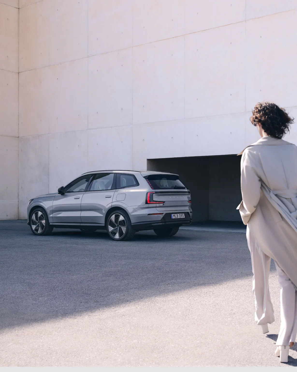 Woman in a long cream coat walking towards a Volvo EX90 parked beside a white building