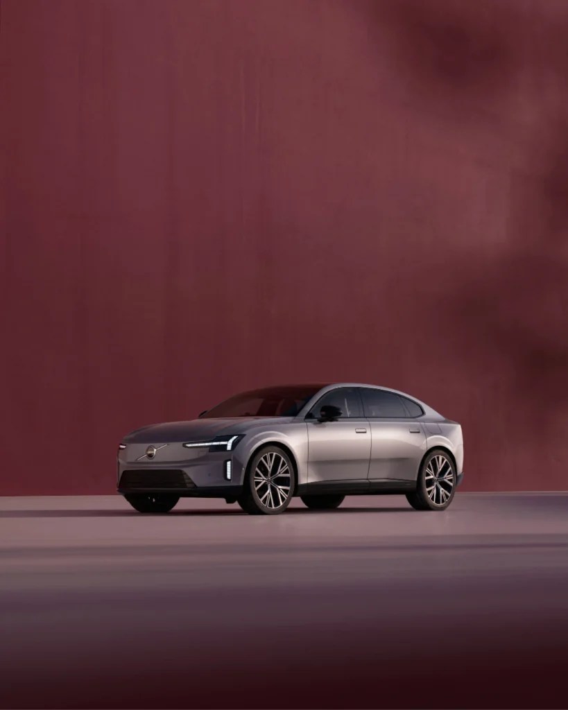 Volvo ES90 electric sedan in a grey metallic finish, parked in front of a dark red wall