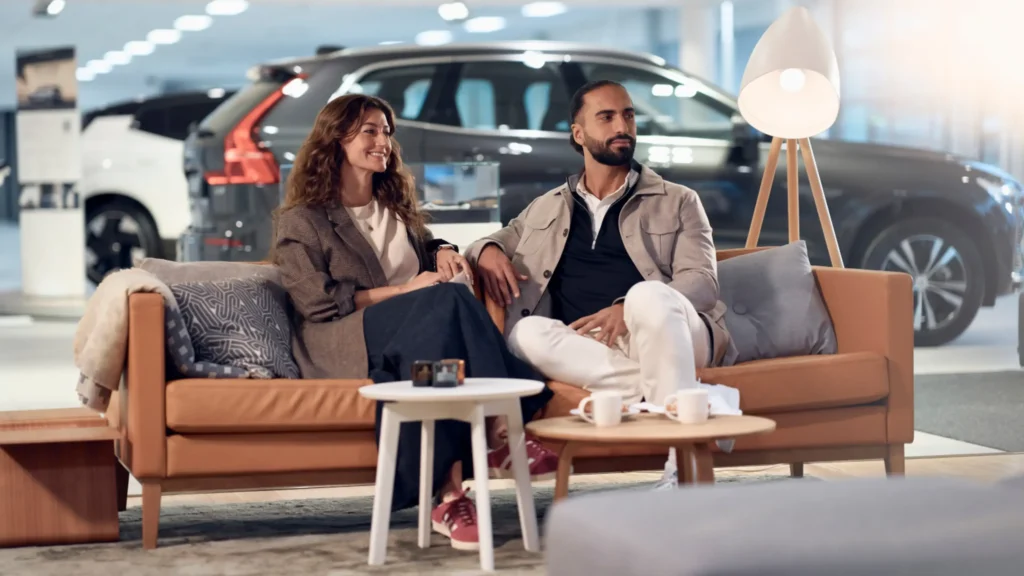 A couple seated on a tan leather sofa in a Volvo showroom, with Volvo SUVs behind them