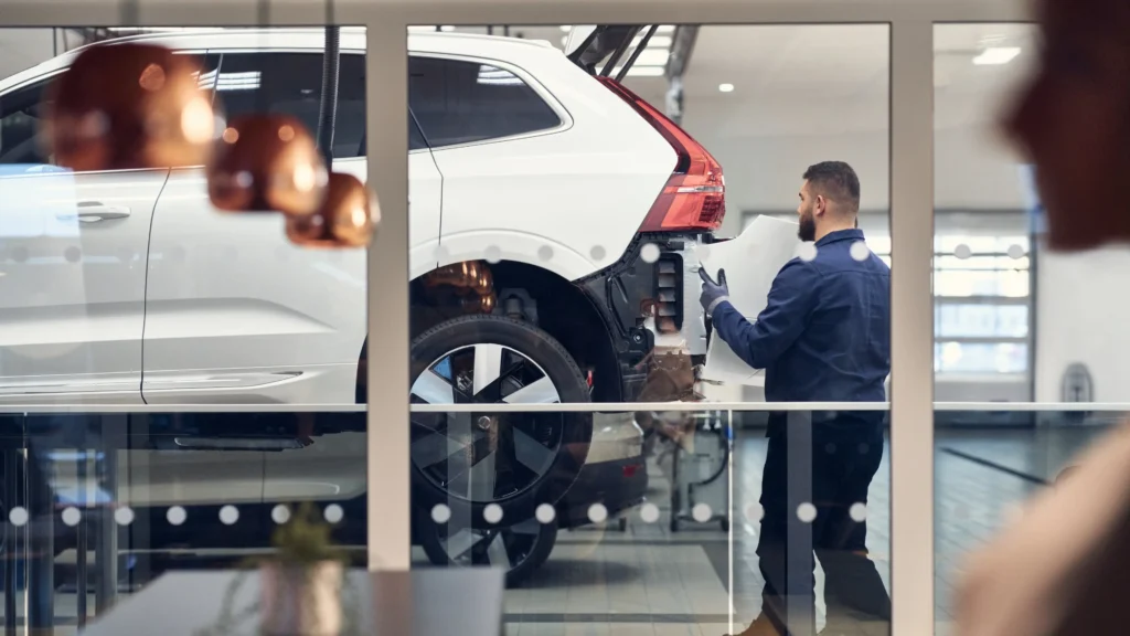 A Volvo service technician beside a white Volvo SUV in a glass-walled service bay