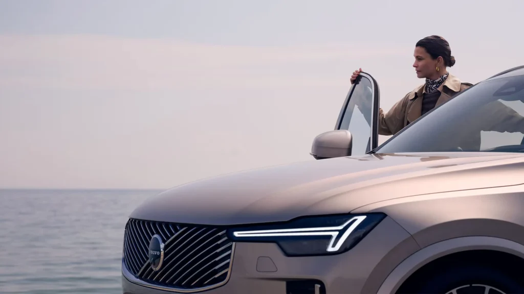 A woman opening the driver’s door of a champagne Volvo SUV parked by the sea