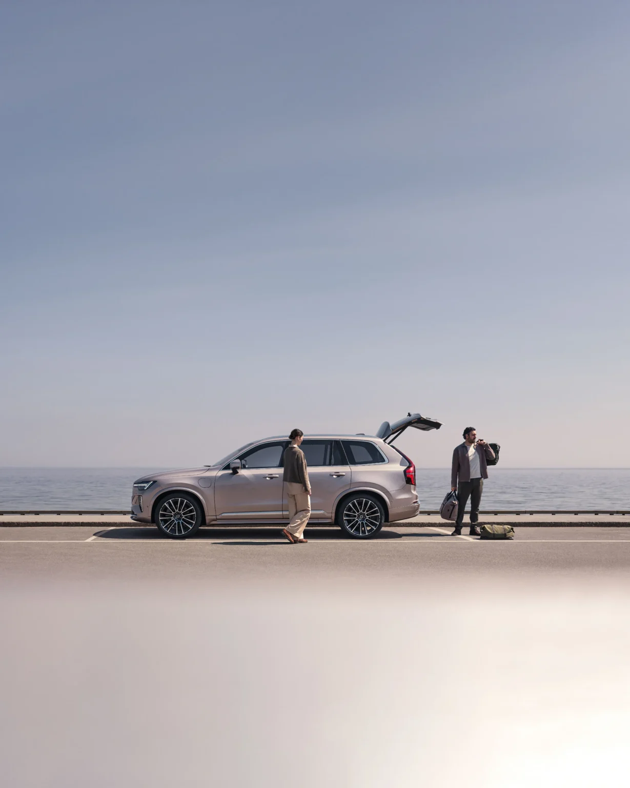 A man and a woman packing their Volvo XC90 on a seafront road, its tailgate open