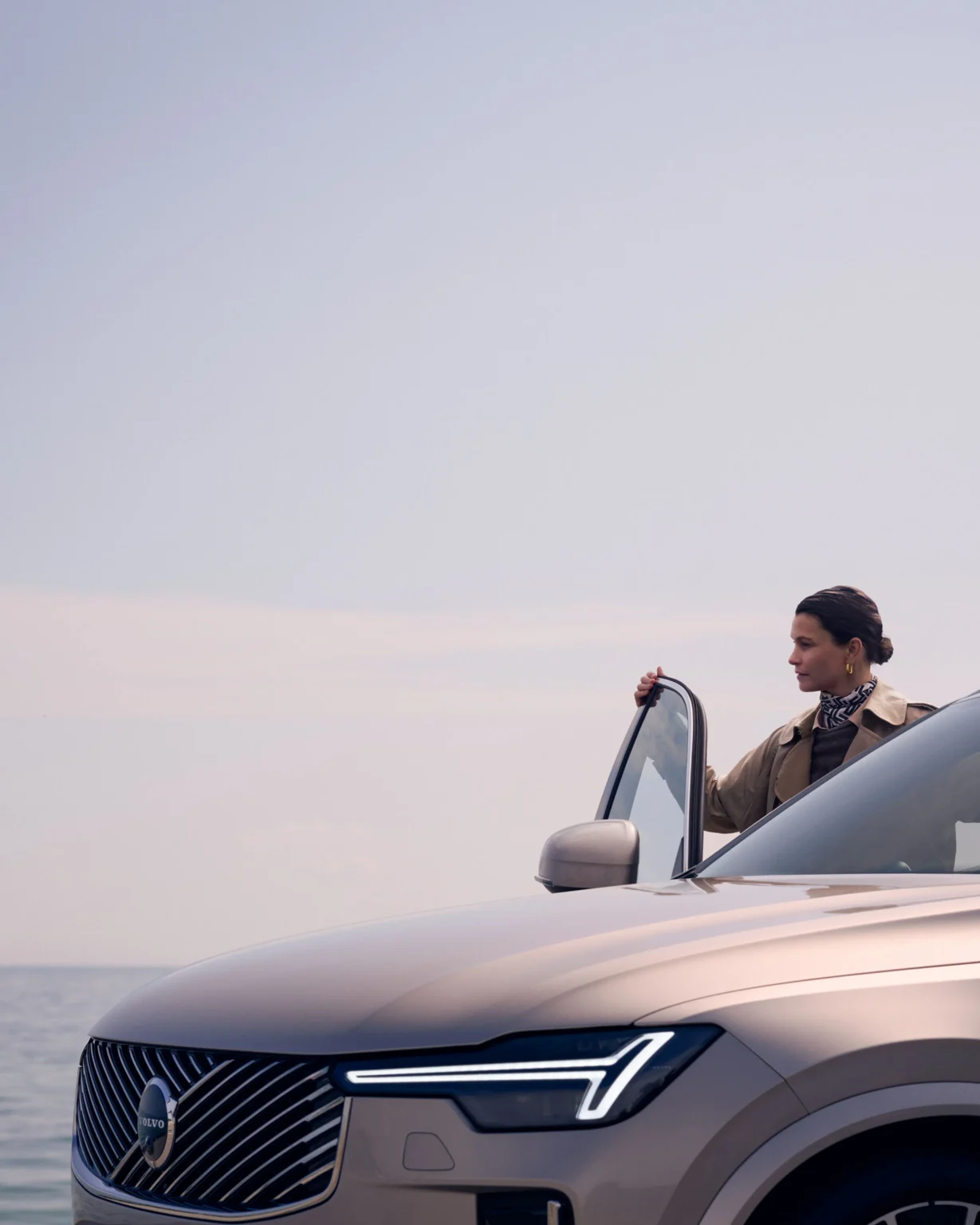 A woman at the open door of a Volvo XC90 by the sea, seen past its headlight and grille
