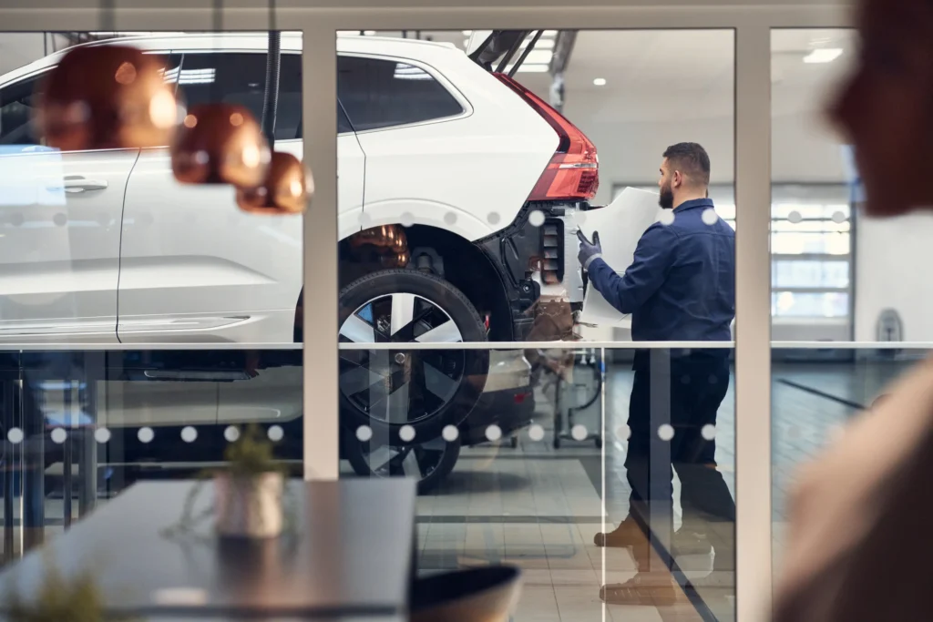 A Volvo technician working beside a white Volvo SUV raised on a lift, seen through a glass wall