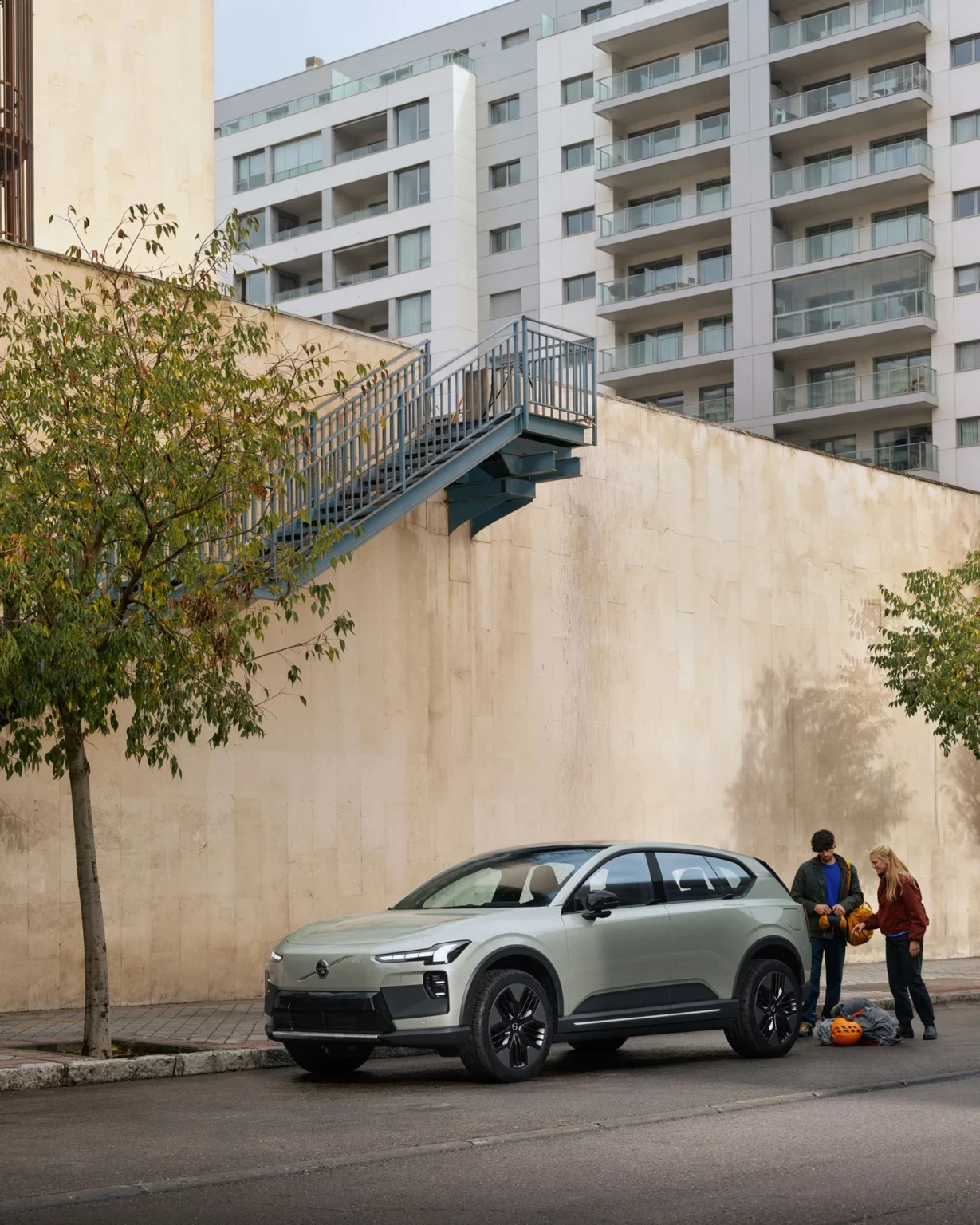 Volvo EX60 Cross Country parked on a city street below an outdoor staircase, two people with bags beside it