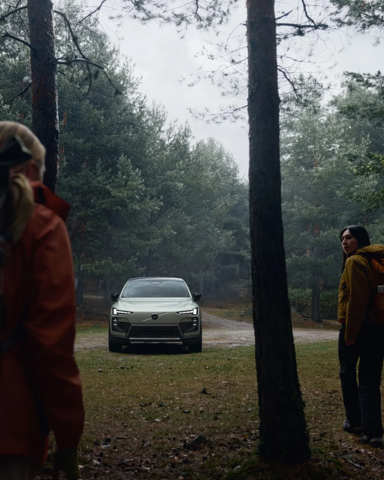 Volvo EX60 Cross Country parked among trees, with two people in outdoor jackets nearby