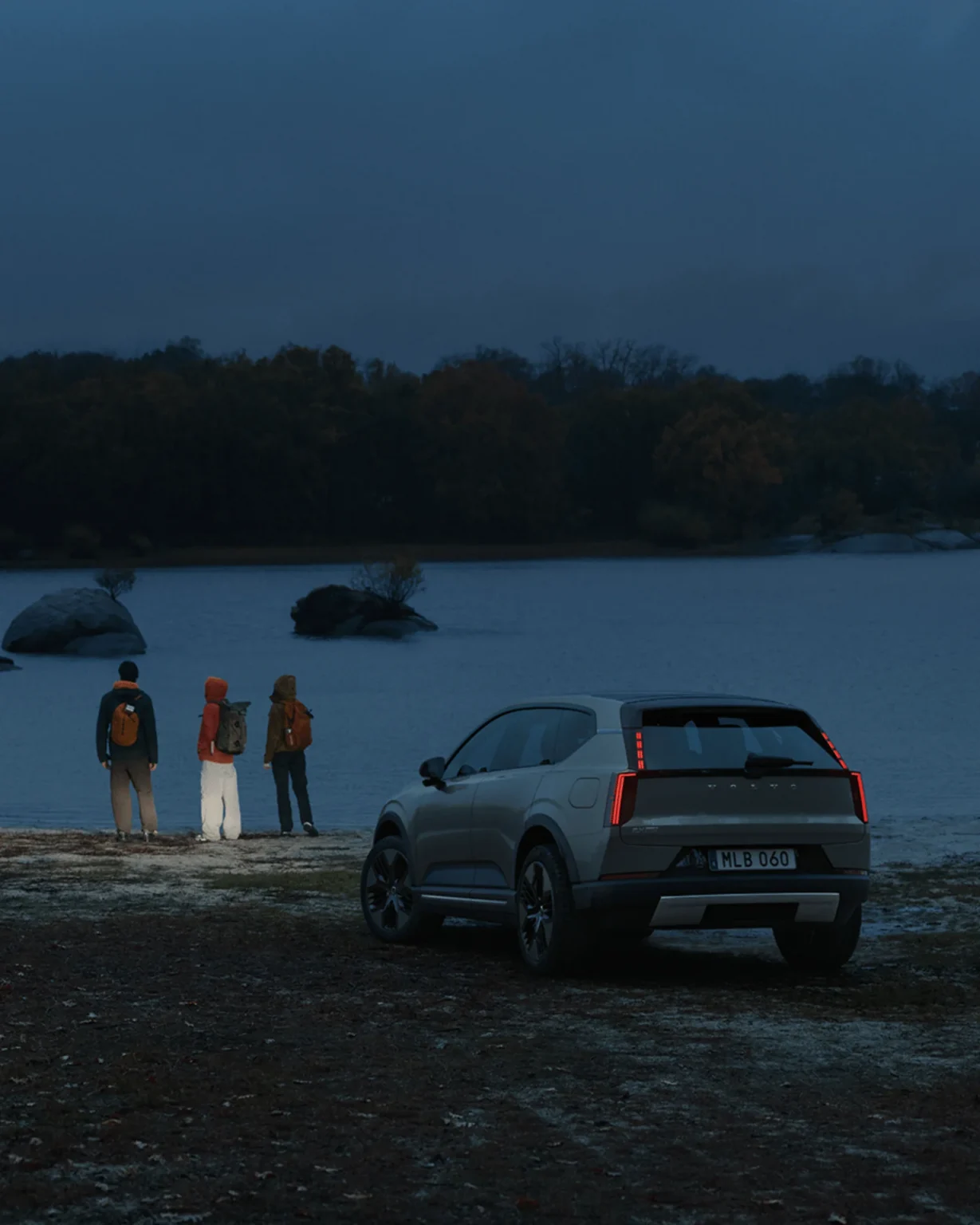 Rear of a Volvo EX60 Cross Country parked by the water at dusk, with three people looking out