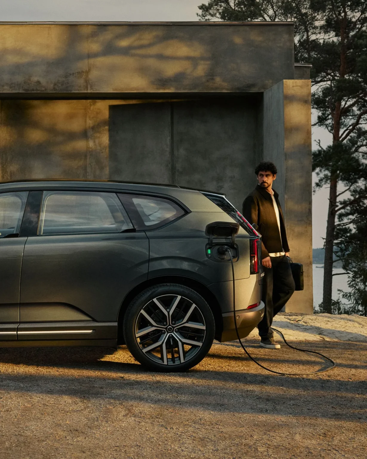 Person standing beside a Volvo EX60 plugged into a home charger at dusk