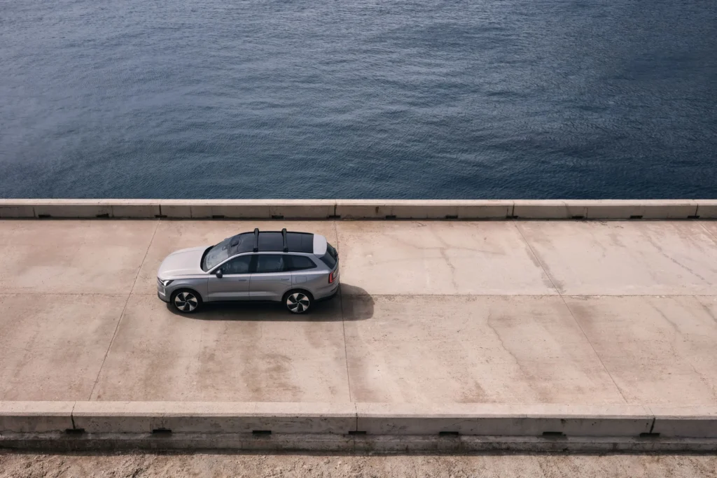 A grey Volvo EX90 with roof bars parked on a concrete pier beside the sea, seen from above
