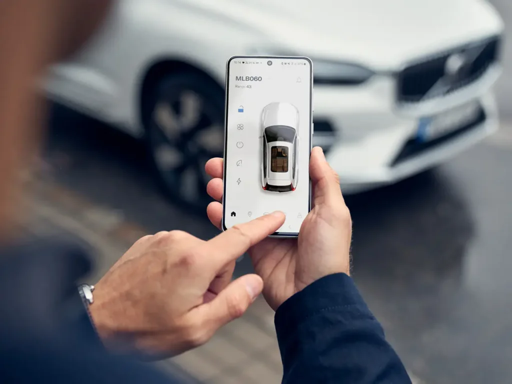 A hand holding a phone showing the Volvo Cars app, a white Volvo behind it