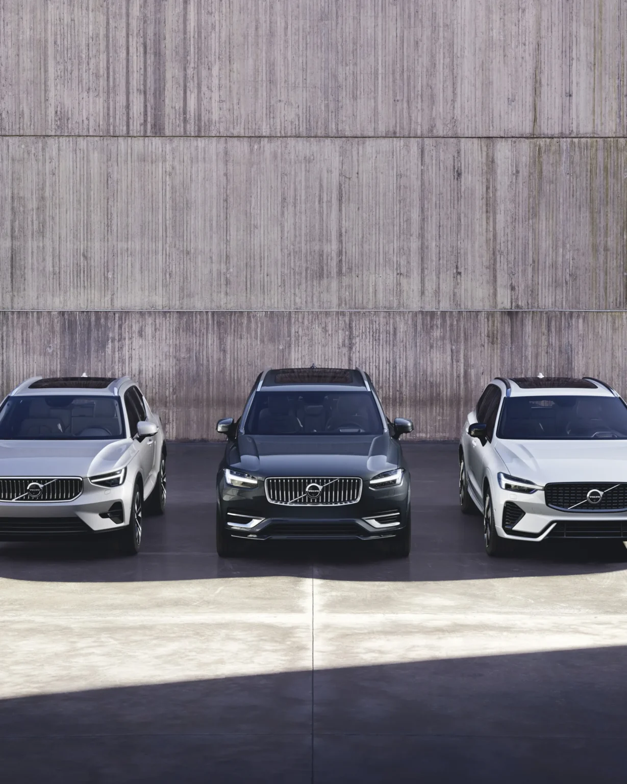 Three Volvo SUVs in silver, dark grey and white parked side by side in front of a concrete wall