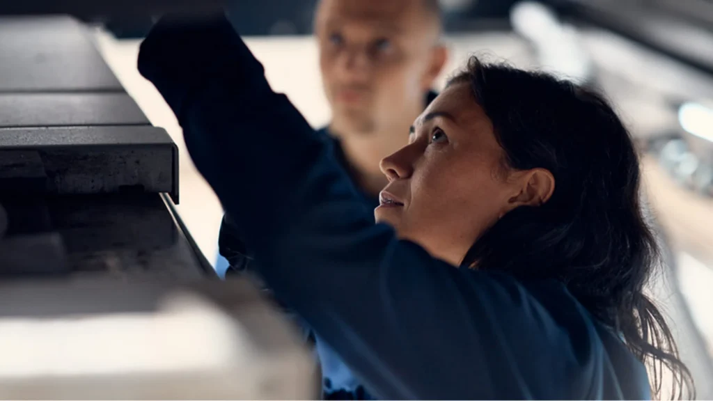 A technician in a navy uniform inspecting the underside of a car on a lift, with a colleague behind her