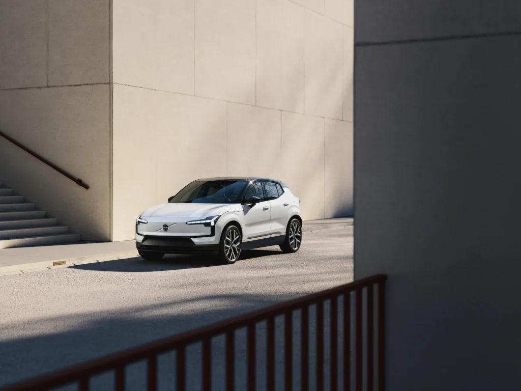 A white Volvo EX30 parked in sunlight between pale concrete walls, behind a red railing