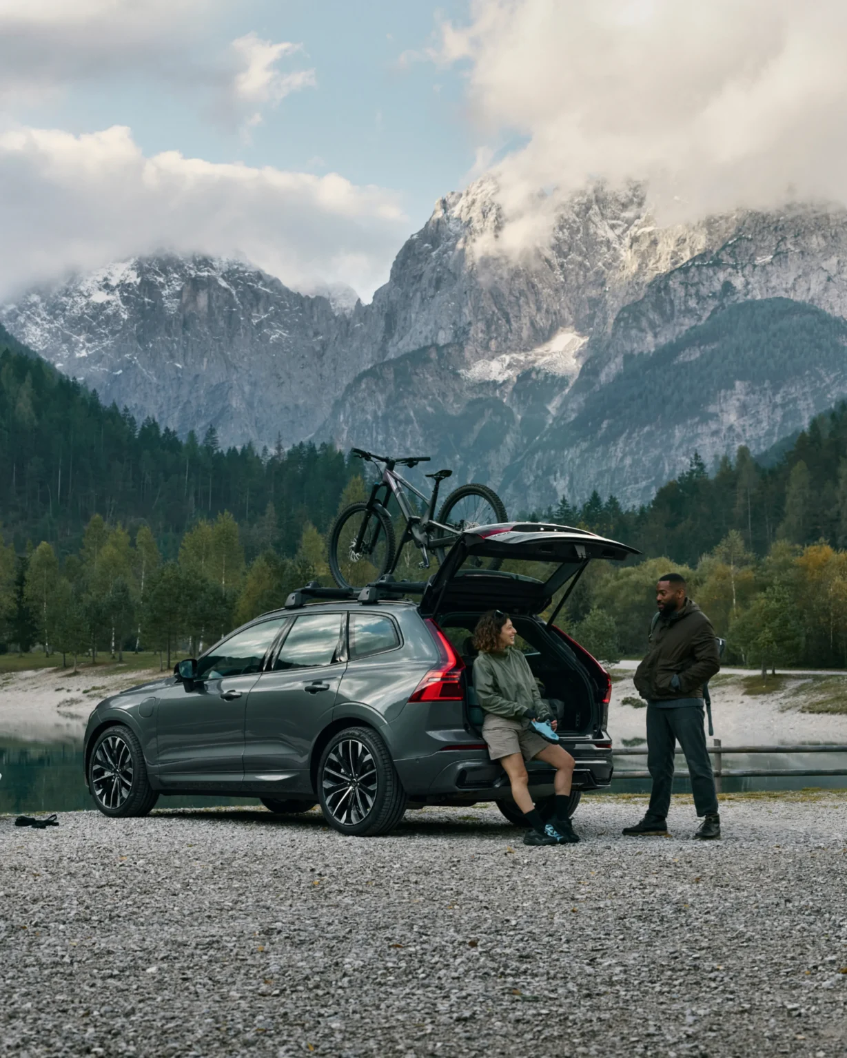 Two people relaxing at the open boot of a Volvo XC60 beside a mountain lake, a bike on the roof