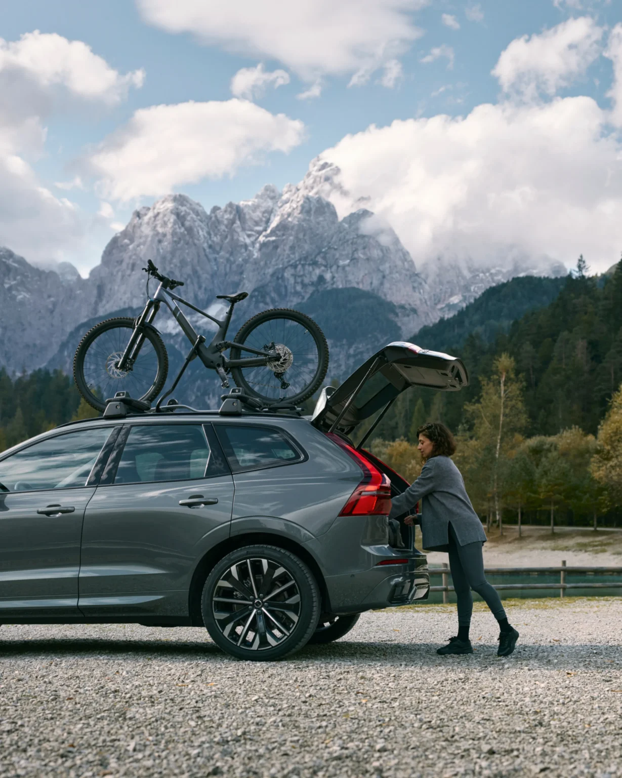 A woman loading the open boot of a Volvo XC60 by a mountain lake, a bike on its roof