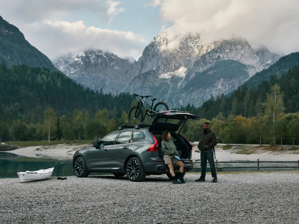 Two people relaxing at the open boot of a Volvo XC60 beside a mountain lake, a bike on the roof
