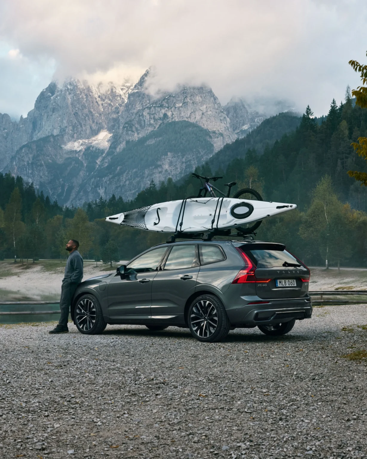 A man standing beside a Volvo XC60 with a kayak on its roof, mountains behind