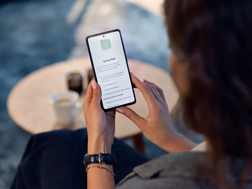 A woman holding a phone showing a Volvo service plan, a coffee cup on the table behind